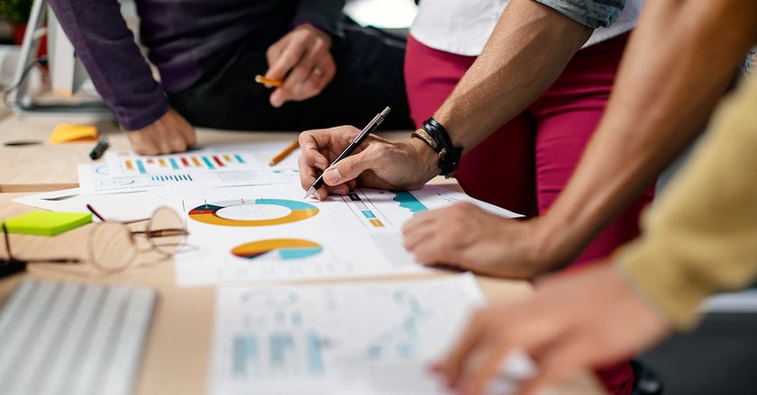 Hands gather around paper graph on table as one person writes with pen