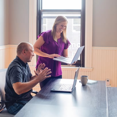 Woman holding laptop smiles while standing next to man sitting at laptop