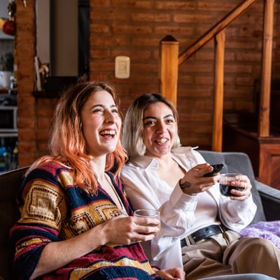 Two girlfiends laugh on cough with wine and laughing while watching tv