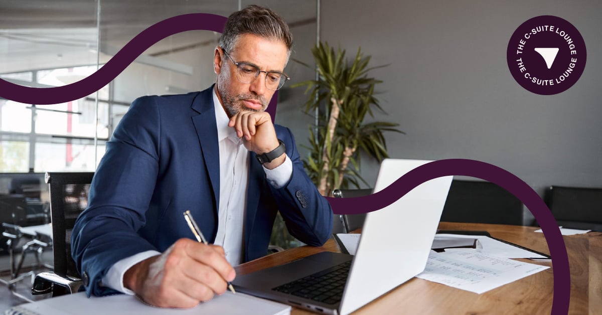 Male CEO in suit sits at desk in front of laptop holding pen 