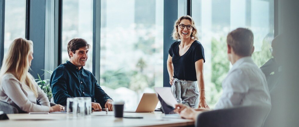 Woman manager stands at conference table while team sits and laughs.
