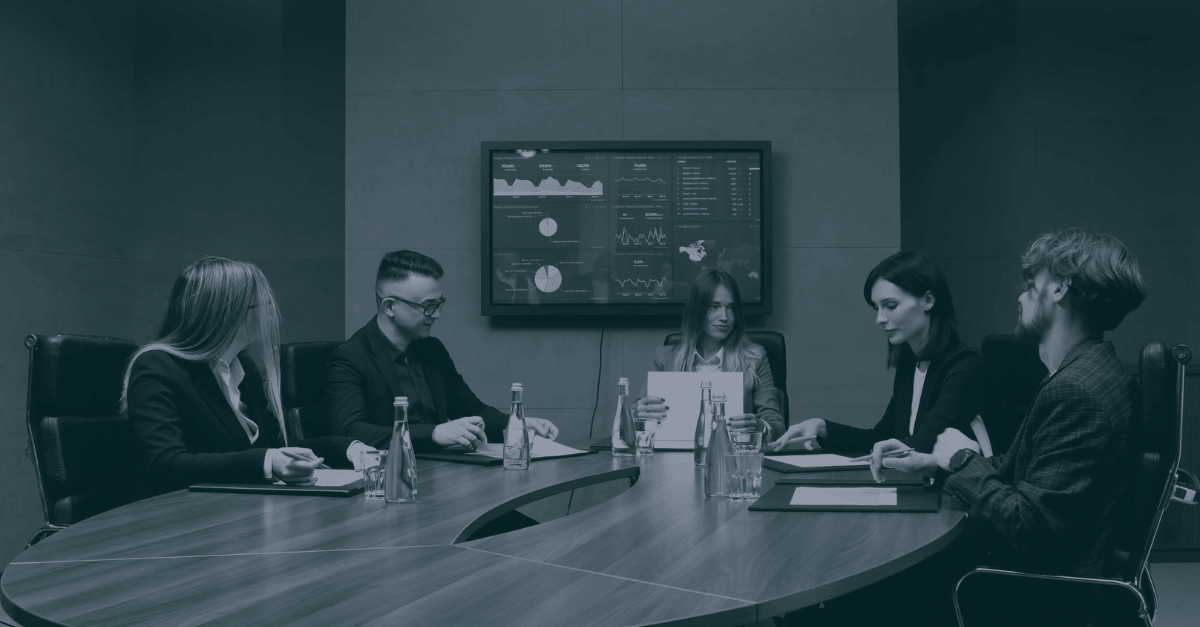 Team of business professionals in a conference room meeting around a table, with a data dashboard displayed on a screen behind them