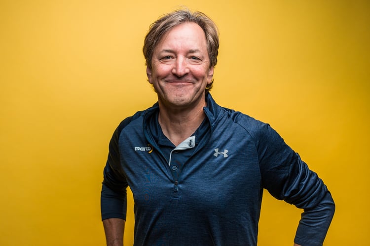 Man in Marco jacket stands in front of yellow background smiling.