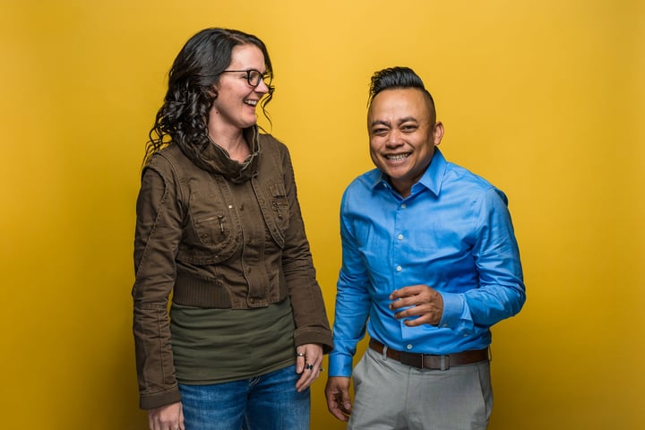 Two people stand laughing in front of Marco yellow background.