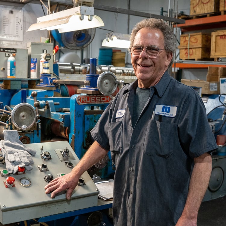 Mead Metals worker wearing branded shirt stands at factory machine smiling.