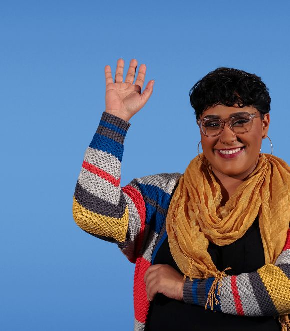 Woman in scarf and sweater raises hand and smiles.