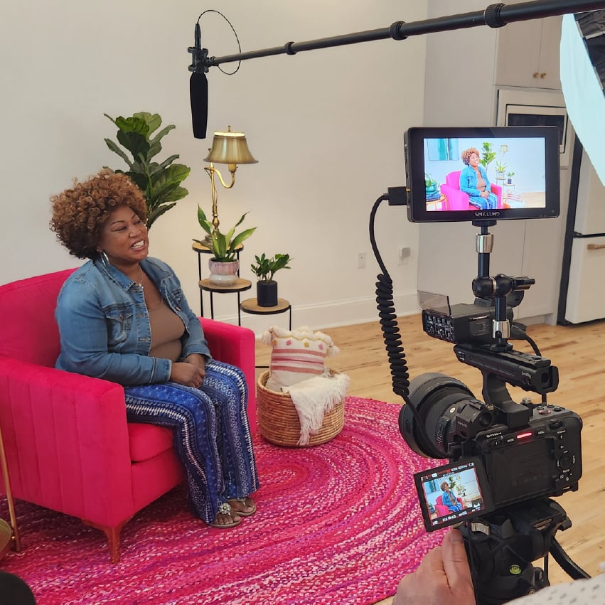 A camera screen shows a woman sitting in a pink chair talking to someone off frame