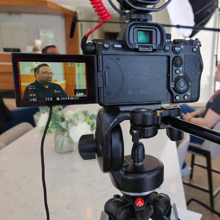 A camera screen shows a man in the background talking to someone off frame