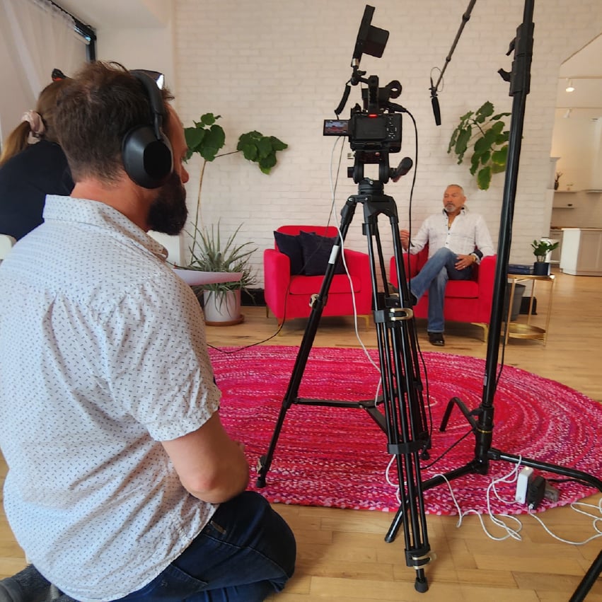 A man with headphones on in front of a camera watches as a man in the background sits and talks